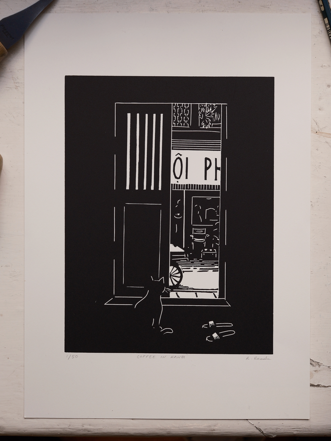 A black and white print depicting a cat watching a street in Vietnam from behind a shutter door. Slippers are by the door and a coffee shop across the road. Done in a simplistic and stylistic way