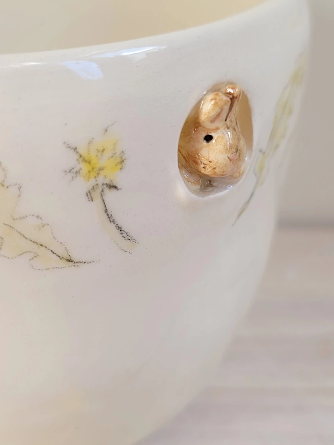 a close up of the head of a miniature beige bunny peeking through a hole in a white pottery cup with hand painted dandelion
