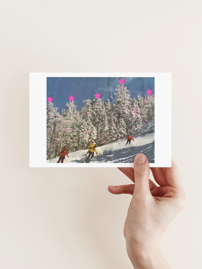 A vibrant Christmas card and brown kraft envelope. The image shows skiers on the tree-lined slopes. The trees are decorated for Christmas.
