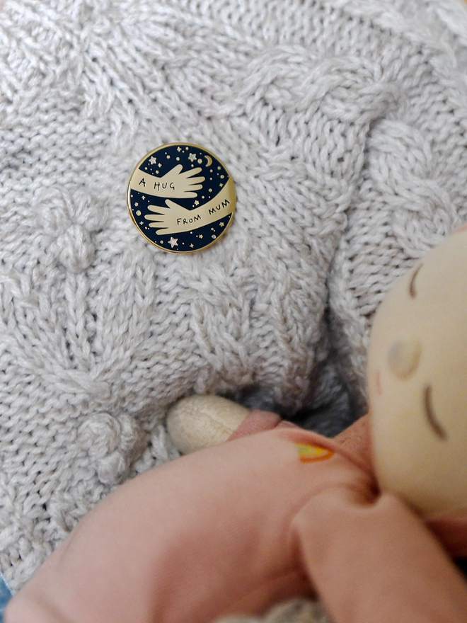 Child wearing a knitted cardigan with a navy blue and gold hug from mum pin attached is holding a baby doll
