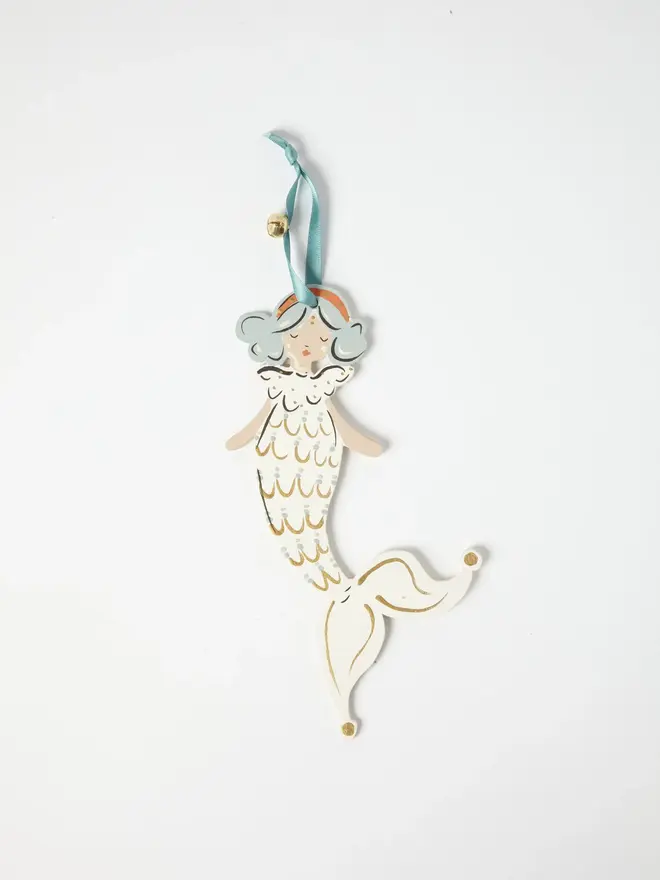 Mermaid Tree Dancer. a flat lay product shot of the mermaid tree dancer hanging ornament in cream and gold tones, on a plain white background. the mermaid figure has pale blue curled hair and an orange headband, dressed in a cream scalloped scale-patterned tail with gold detailing, curling into a jester-style pointed tip with gold-tipped ends. the figure hangs from a teal ribbon with a small gold bell attached, giving the ornament a festive, folk-art charm.