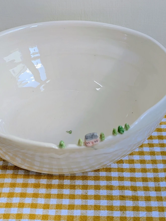 Handmade ceramic white bowl with a tiny pink house and tree details on the rim on a yellow gingham table cloth