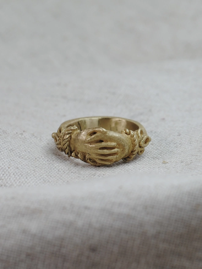 The image of a gold toned brass ring with 2 right hands holding each other and details on the shoulders of the ring. The ring is on a piece of natural linen 