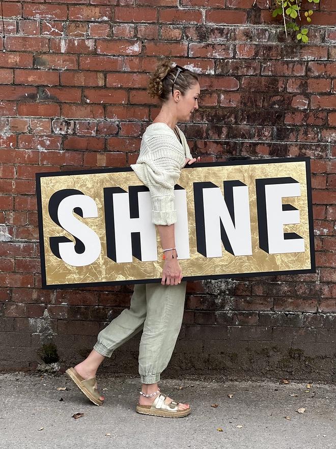 A person waking past a red brick wall holding a large wooden sign reading SHINE on a gold foil background 