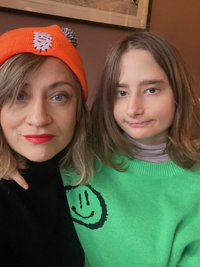Piper has her arm around her mum Kate who is wearing an orange bobble hat, black top and bright red lipstick and piper has green jumper with a black smiley face. they are both looking straight at the camera