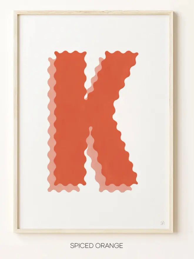 Framed wall art print featuring a large decorative letter “k” in soft orange tones with a ric rac–style wavy edge design, centered on a white background. 