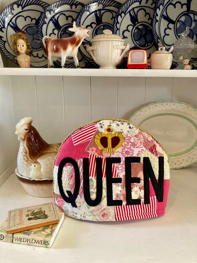 Fun, Queen tea cosy on a dresser with crocery.