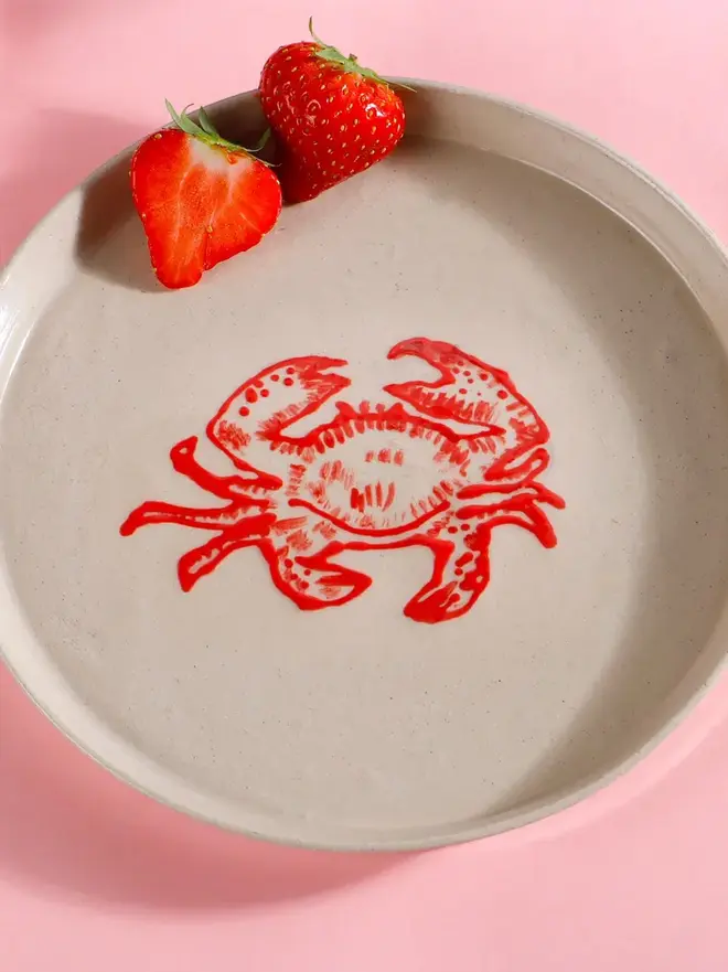 Handmade Ceramic Side Plate With A Red Crab Design, a white side plate with a red crab on a pink table. 