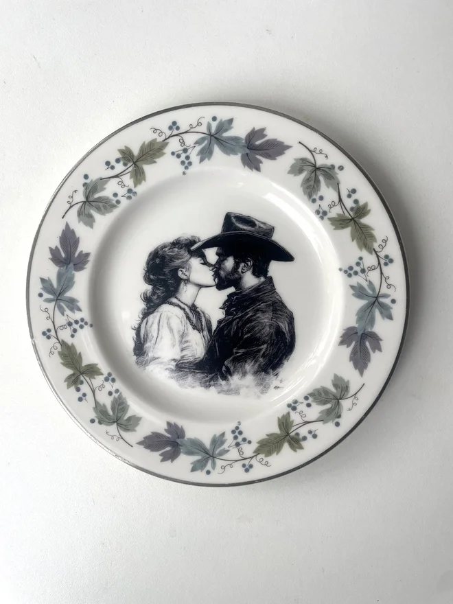 a vintage plate with an ornate border has a black and white vintage illustration of a cowboy and cowgirl sharing a kiss in the centre