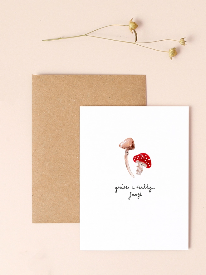 An image of a mini, rectangular white greetings card with an illustration of a couple of red and white toadstools with smiley faces. A hand written 'you're a really fungi' message is seen under the central illustration. The mini greetings card is placed on top of a rectangular brown kraft envelope.