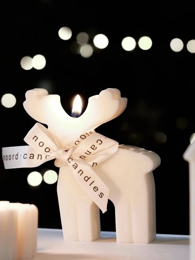 Reindeer Sculpture Candle. A lit cream reindeer candle tied with a Noord Candles ribbon bow, glowing against twinkling bokeh lights beside a little house candle. The sculptural festive candle makes a magical centrepiece for Christmas evenings.