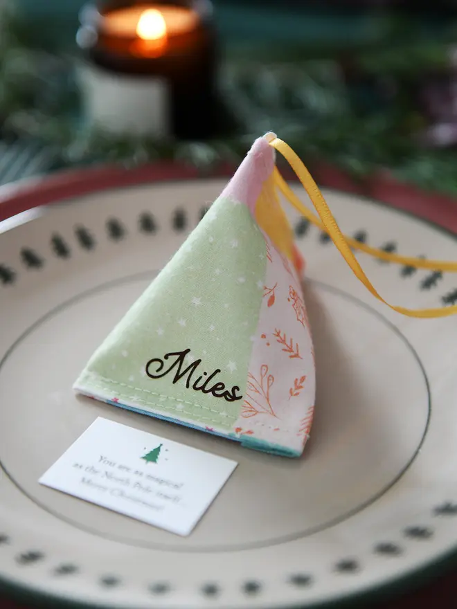 A pastel patchwork fabric fortune cookie with gold personalised name “Martha” sits on a Christmas dinner plate, ready as a handmade festive place setting with a hidden Christmas wish card beneath.