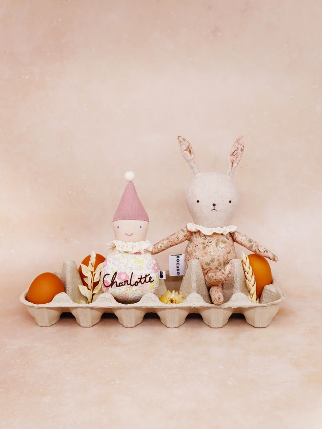 Handmade linen bunny doll in pink floral outfit and frilly lace colour, sitting in an egg carton with eggs.