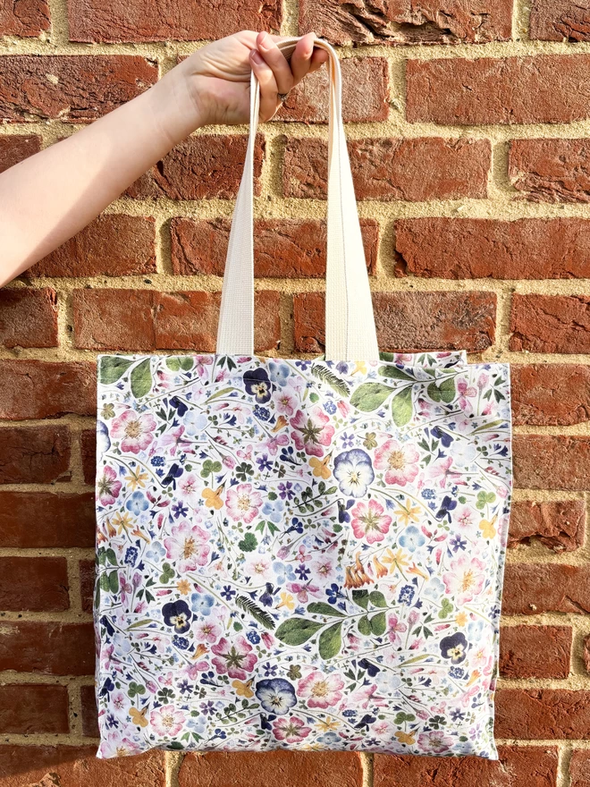 Pretty pressed flower shopping bag being held against a brick wall backdrop. With strong handles. The perfect gift for nature lovers.