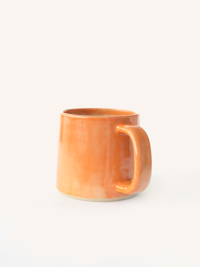 A handmade ceramic standard mug in glossy mandarin orange glaze with a chunky loop handle, photographed against a plain light grey background