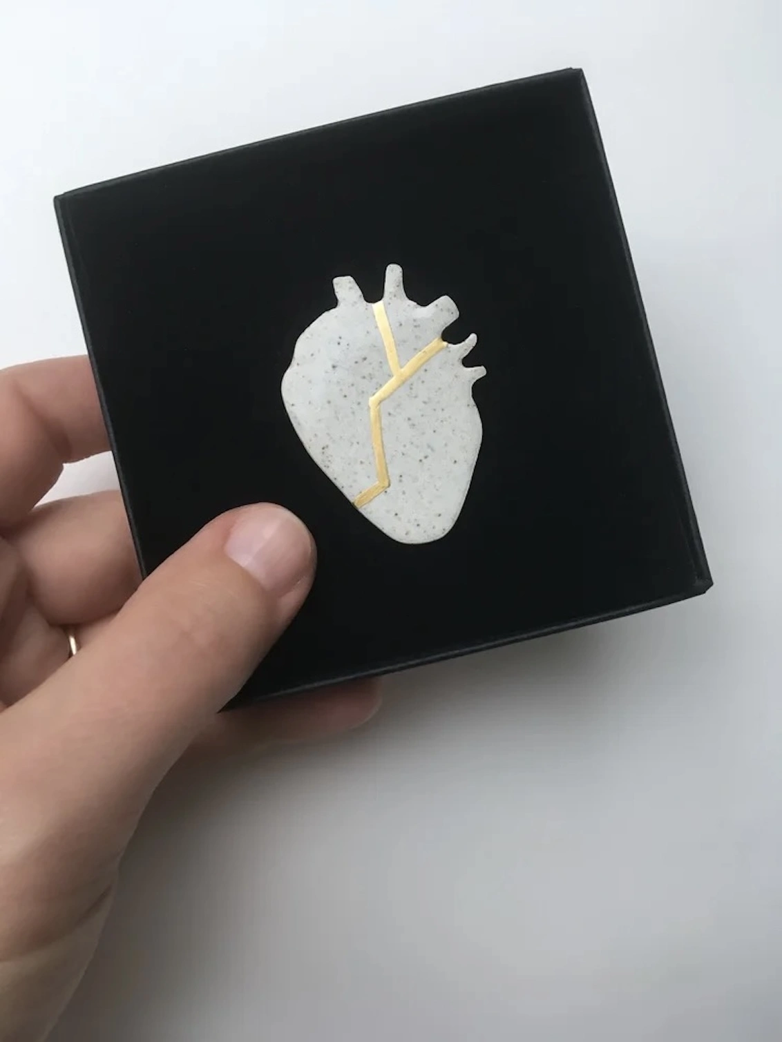 (Un)Broken Heart Pin, a white and golden heart pin sitting on a white surface in a black box. 