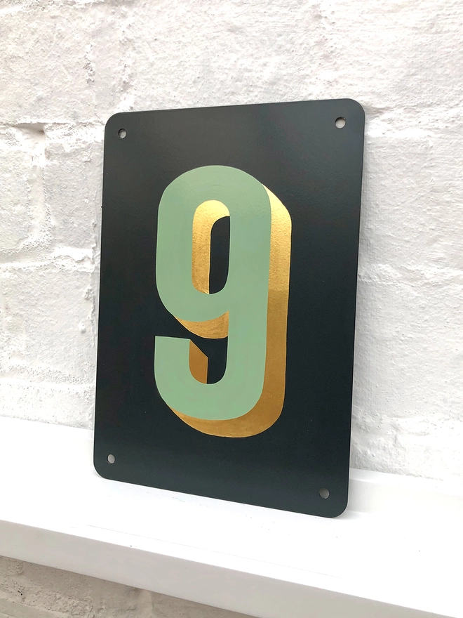 Sage green and gold leaf house number 9, on anthracite grey metal plaque, against a white brick wall.