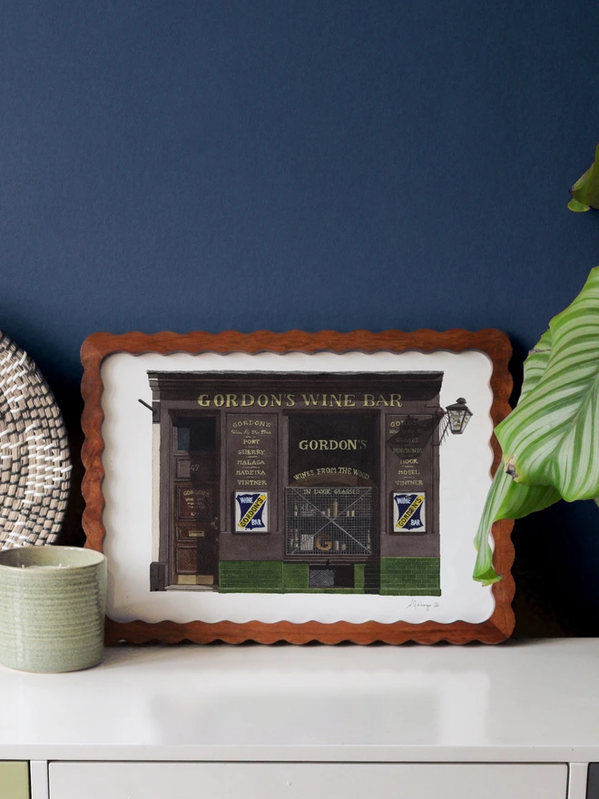 A framed watercolor print of Gordon's Wine Bar displayed against a dark blue wall beside a woven tray and a large green leaf.