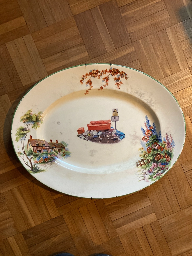 large oval vintage plate featuring a cottage on the left with a floral border. In the centre sits a pile of rubbish with an old sofa on top, next to a sign stating 'no fly tipping'.
