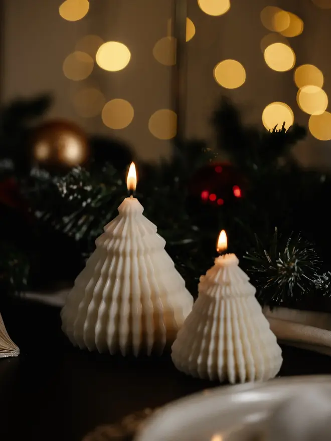 Origami Christmas Tree Candles (Set Of 2). Two lit cream cone tree candles with intricate fluted ridges, glowing beside frosted pine garland and a paper ornament on a dark table. The textured sculptural design gives a soft, cosy glow to winter evenings.
