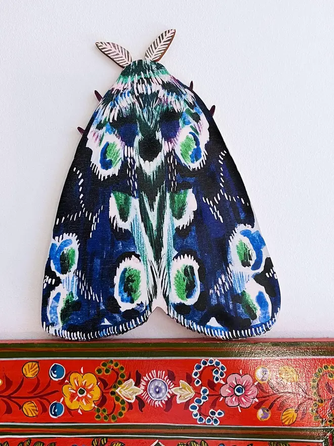 large wooden moth wall decoration. large blue wooden moth decoration hung on plain white wall.