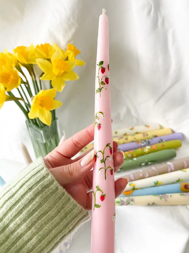 Wild Strawberry Pink Hand Painted Candle, a woman holding the pink candle against a white backdrop accompanied by a flower. 