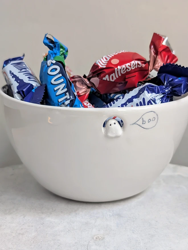 small white ceramic bowl with a tiny Halloween ghost and the word boo in a speech bubble the dish is full of chocolate sweets