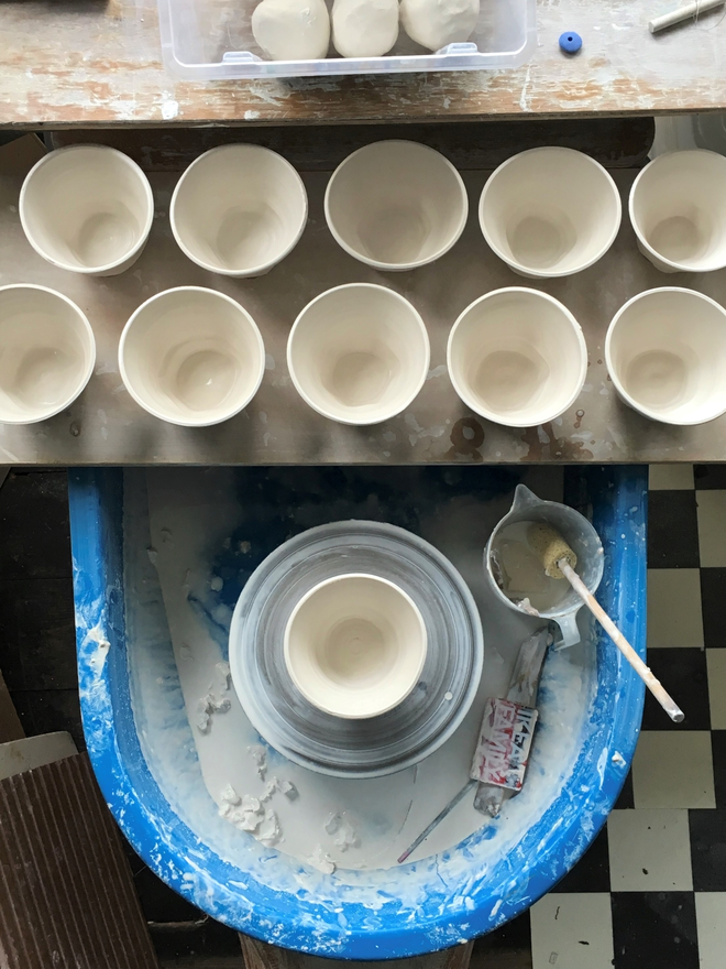 bird view of a blue potters wheel  with 11 freshly thrown porcelain cups , the wheel tray is covered in slippert white porcelain slip