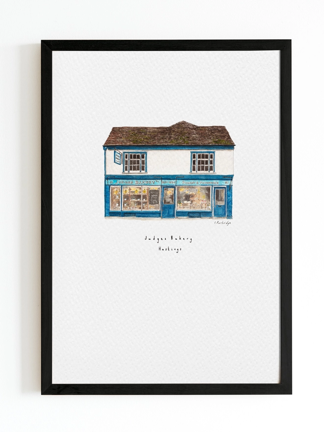 Watercolour illustration of Judges Bakery a beautiful blue shopfront with windows full of bakery goods sits below a white first floor and dark brown / grey roof. The illustration is a small watercolour painting in the centre of the white page and sits within a black frame. 