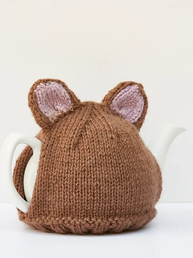 Brown Bear Tea Cosy Teddy Bears Picnic Treat, a brown bear tea pot cover sitting on a white tea pot, on a white surface against a grey backdrop. 