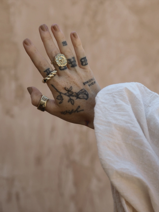 The image of a tattooed female hand wearing a hand carved gold toned brass sun face ring, a twisted gold toned brass ring with a blue sapphire and a thick cigar style gold toned brass ring with star set crystals in front of a rustic terracotta coloured plaster wall