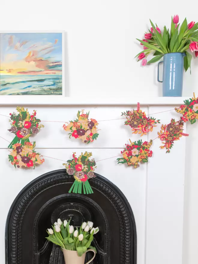 Wide shot of complete red bouquet garland draped across white fireplace, white tulips, blue vase and beach scene canvas