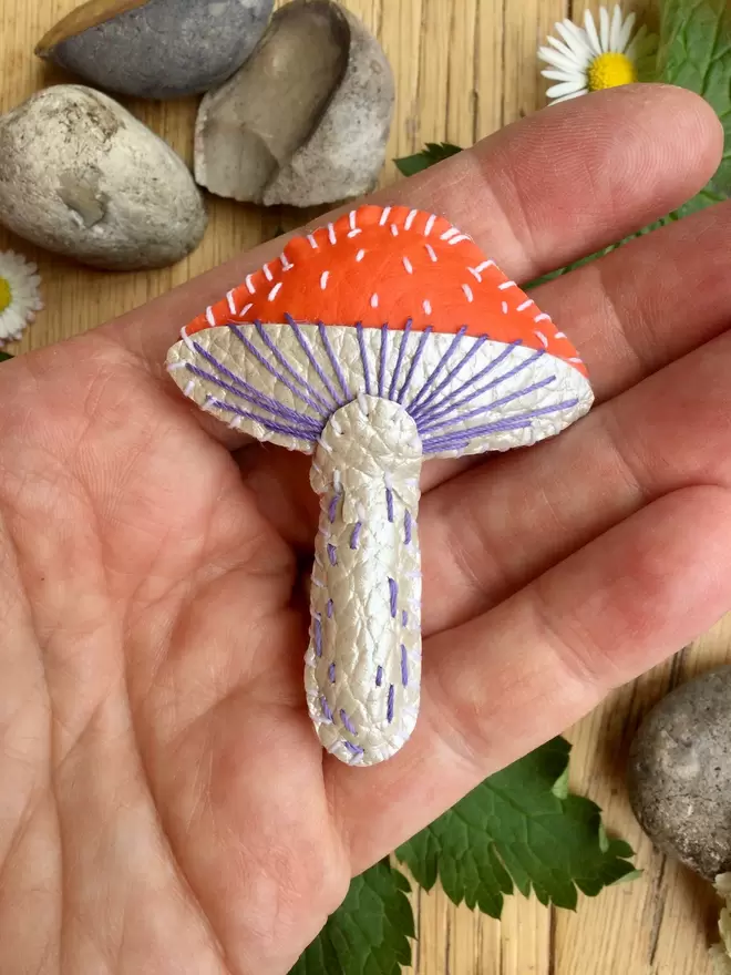 The palm of a hand, holding an orange and white faux leather stitched toadstool brooch 