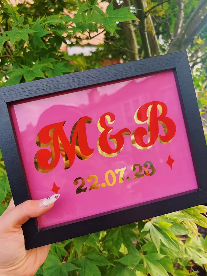'M & B' wedding sign with a curly script style on pink background, with red lettering and gold leaf shade and date.