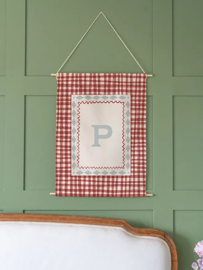 Personalised Gingham Initial Fabric Banner, red hanging on a green wall. 