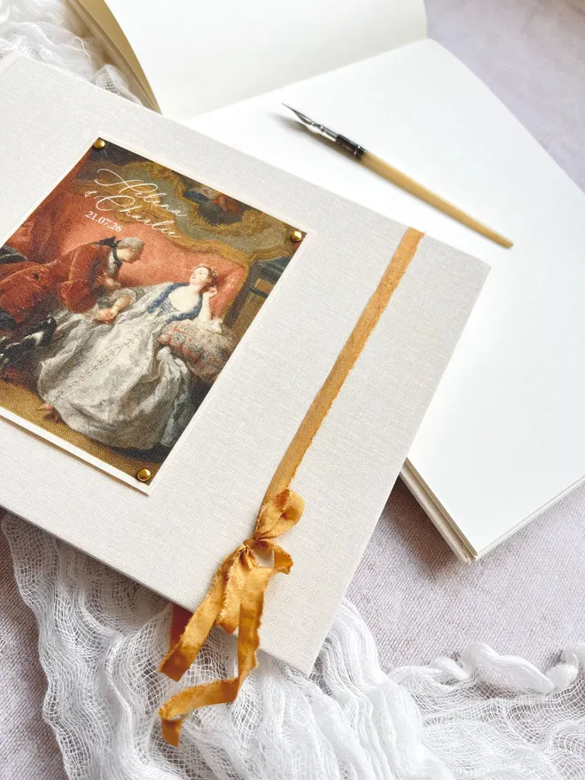 wedding guest book showing the cover and inside pages. The cover is a romantic 18th century rococo scene of lovers with your names and wedding date featured and gold faux silk ribbon. The pages inside are high quality blank ivory writing pages.