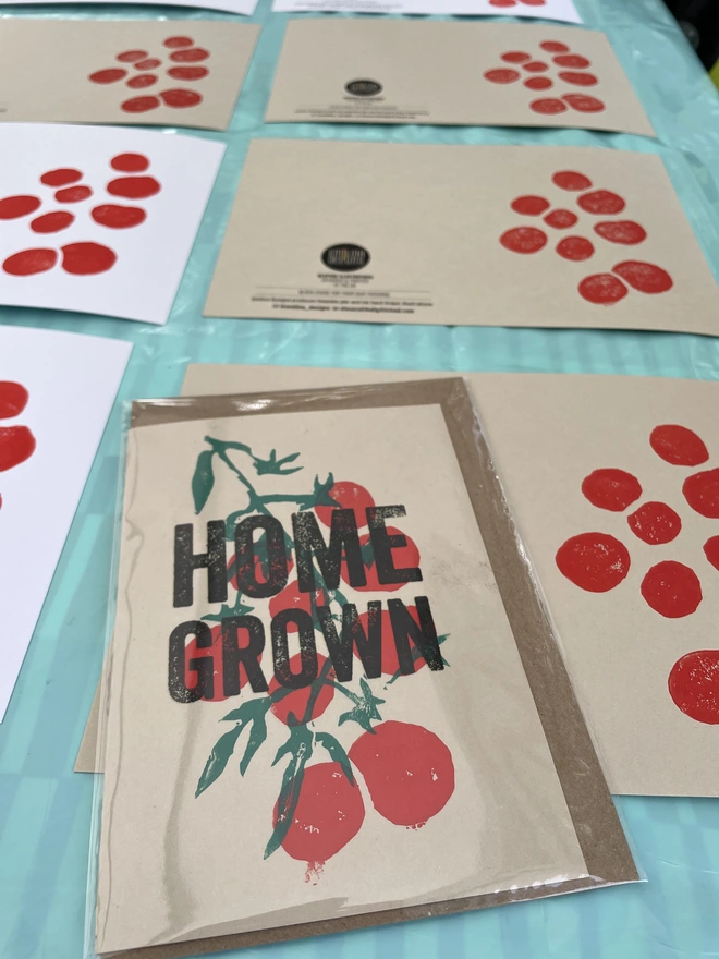 'Home Grown' Tomatoes Lino Garden Print. A display of handmade tomato-themed cards arranged on a table covered with a light blue sheet.