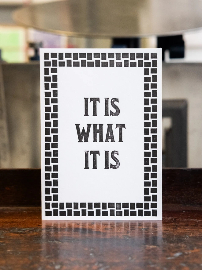 Letterpress card with text design "It is what it is," Surrounded by art deco style border of woven squares.