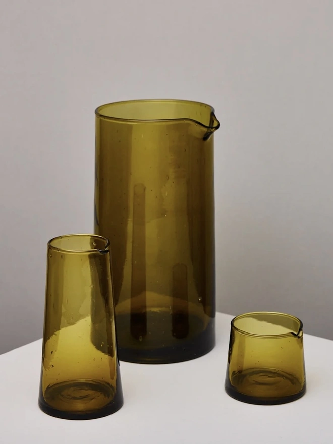 Tall Jug, a tall amber glass jug sitting on a plain counter top against a white backdrop. It is accompanied by other glass drinkware items. 