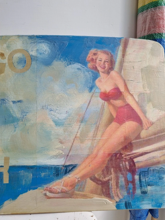 Upcycled art panel in blues and creams featuring a seascape and an image of a retro pin-up lady on a boat and the text reads ‘let’s go to the beach’ in a distressed, shabby chic style