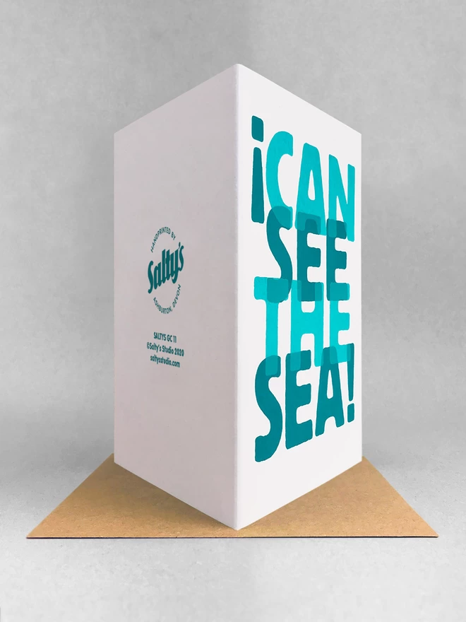 Rear view of I can see the sea, screenprinted words overlapped in two turquoise blues, on a white card. It is stood on a kraft envelope in a light grey studio set.