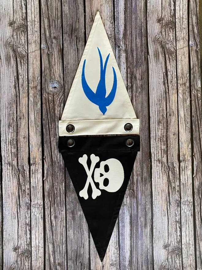 two pennant flags laying on a wooden floor. one is ivory with a blue swallow and one is black with an ivory skull and crossbones
