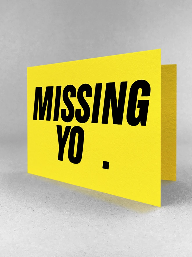 A side on view of a Bright yellow card with the words Missing you and a full stop screenprinted in black. There is a gap where the letter u shoud beissing