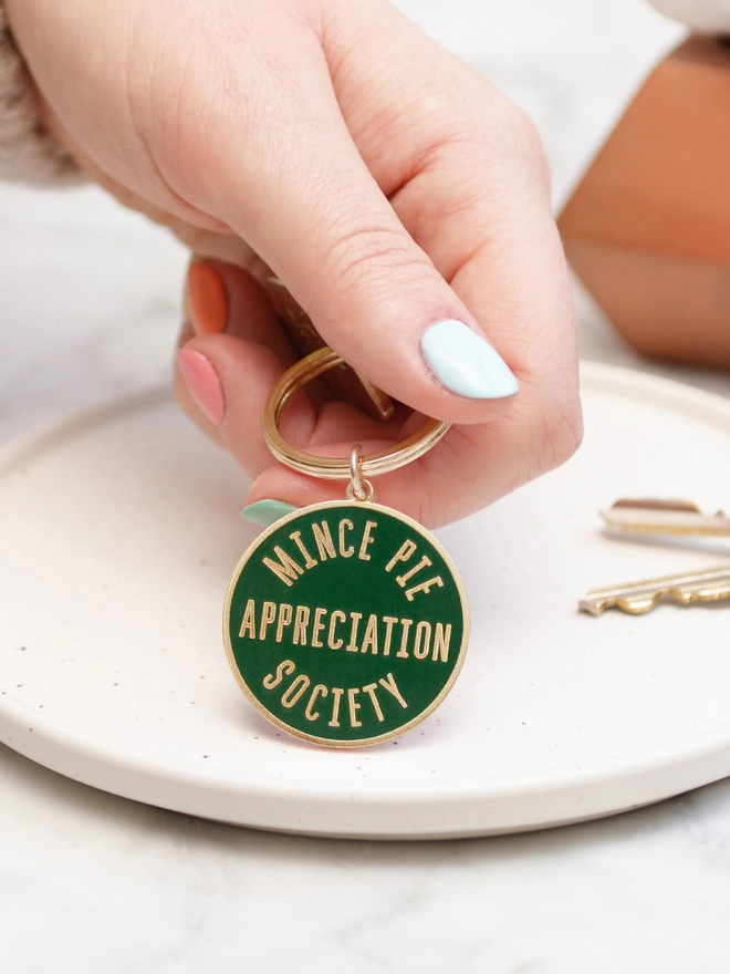 Hand holding out a Green enamel keyring featuring 'Mince Pie Appreciation Society' design