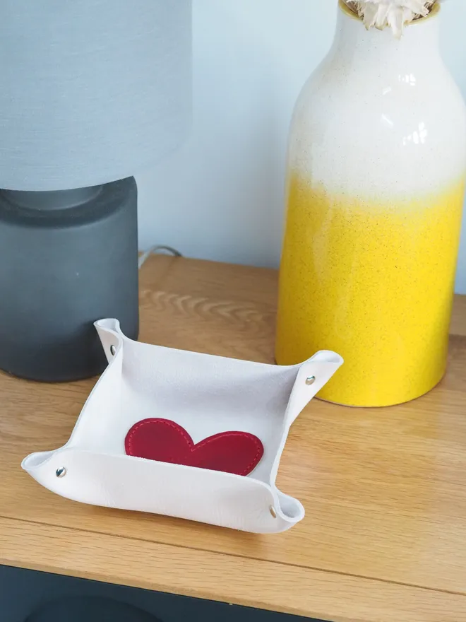 Personalised leather heart coin tray. A natural cream square leather tray with a red leather heart stitched to the base. The heart is personalised with a couples initials and their wedding date. The tray sits on a side table beside a lamp and a vase.