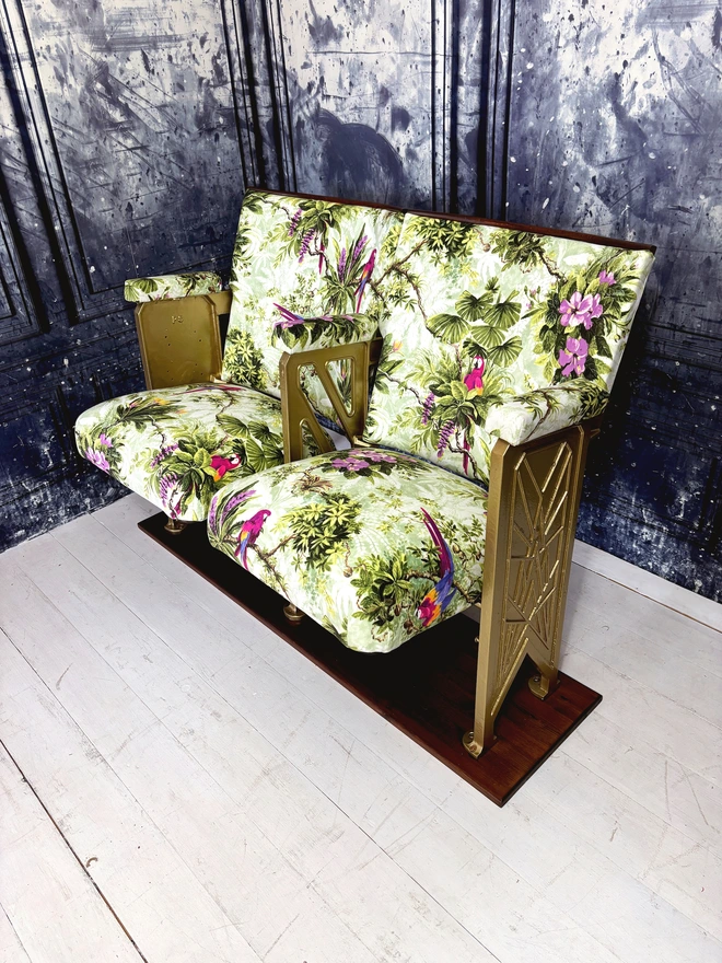 set of two vintage cinema seats upholstered in a purple and green macaws velvet.  Seats are sat on a white wooden floor with a marbled blue wall behind them