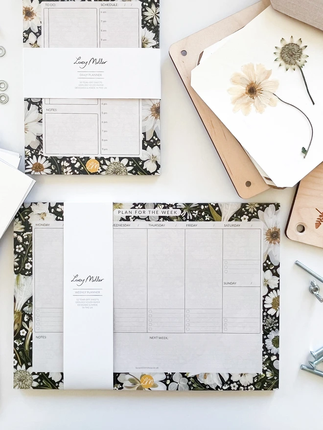 Weekly planner desk pad with pressed white flowers on black background, showing weekday and weekend columns, 'Notes,' and 'Next Week' sections; perfect for busy professionals, students, or home organisation.