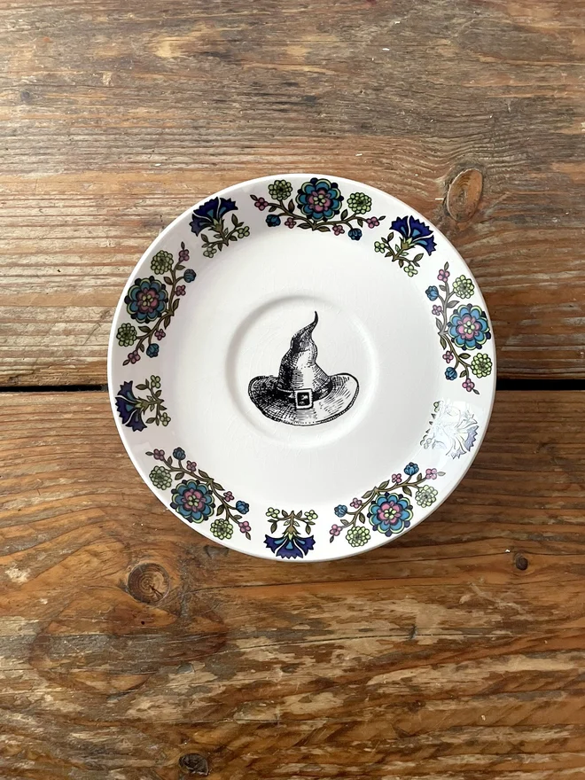 A white plate with an ornate floral 70s border in blues and greens, in the middle is a vintage black and white illustration of a witch's hat