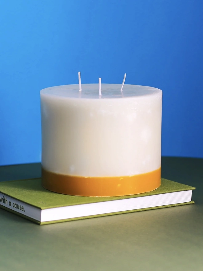 Jasmine, Green Tea & Mulberry Candle (Set Of 1 Or 3 Wicks). the jasmine, green tea and mulberry candle sits on a green book against a bright blue background, showing its three wicks and cream wax with a golden yellow base. the two-tone colouring and textured, slightly uneven wax surface give the candle a rustic, hand-poured finish. the bold blue background highlights the candle's warm, honeyed tones.