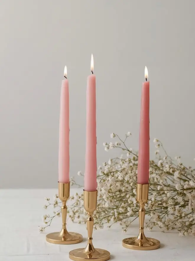 three lit pink dinner candles, sitting in gold candle holders on a white table, with white flowers in the background